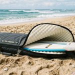 A foam padded surfboard bag lying on a sandy beach with a surfboard partially inserted, showing thick cushioned interior lining