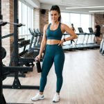 A woman in a well-coordinated fitness outfit including high-waisted leggings and a sports bra, standing in a bright modern gym