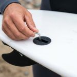 Close-up of a surfer installing a fin screw into an FCS plug on the bottom of a white surfboard