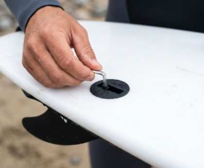 Close-up of a surfer installing a fin screw into an FCS plug on the bottom of a white surfboard