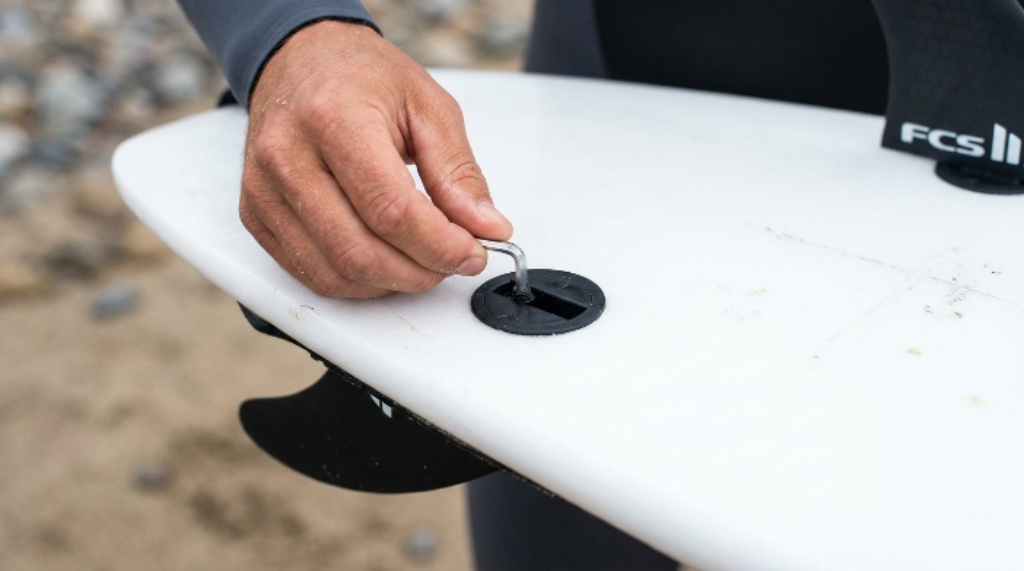 Close-up of a surfer installing a fin screw into an FCS plug on the bottom of a white surfboard