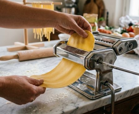 Hands feeding fresh dough through a stainless steel pasta maker.