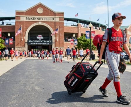 Youth player pulling a wheeled baseball bag toward a sunny diamond.